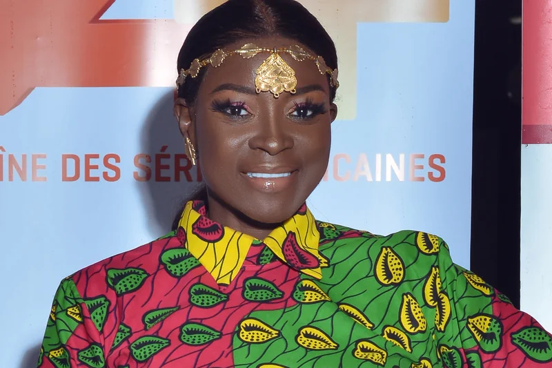 Lucie Memba souriante avec bijou frontal dore et chemise wax rouge, verte et jaune