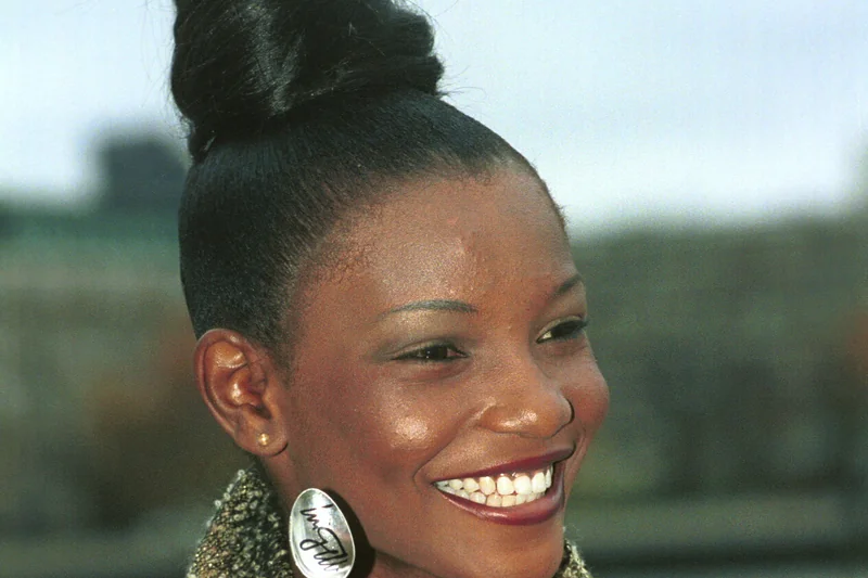 Agbani Darego souriante, chignon haut et veste doree texturee, lors d'un evenement Miss World