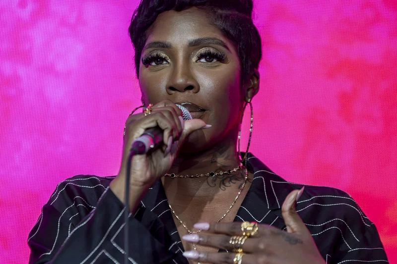 Tiwa Savage sur fond rose avec micro, chemise noire graphique et bijoux dores