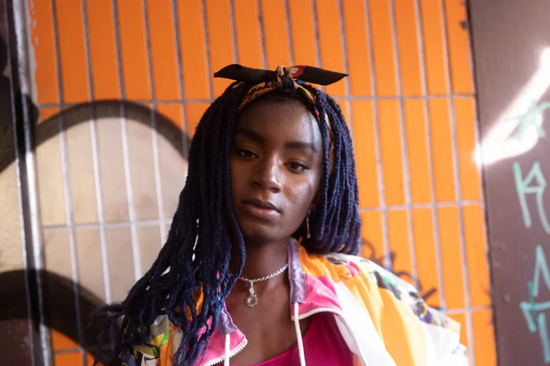 Oumi Janta en robe rose et veste wax colorée devant un fond urbain orange